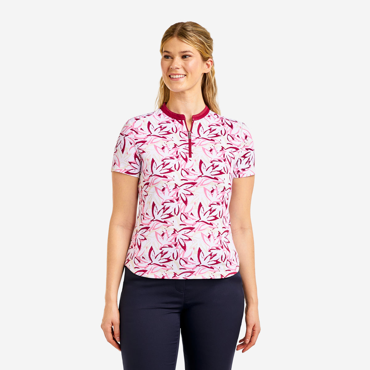 A woman modeling a short-sleeve golf polo shirt with a pink and white floral pattern and a dark pink collar and zipper placket, paired with dark navy pants, against a light gray background.