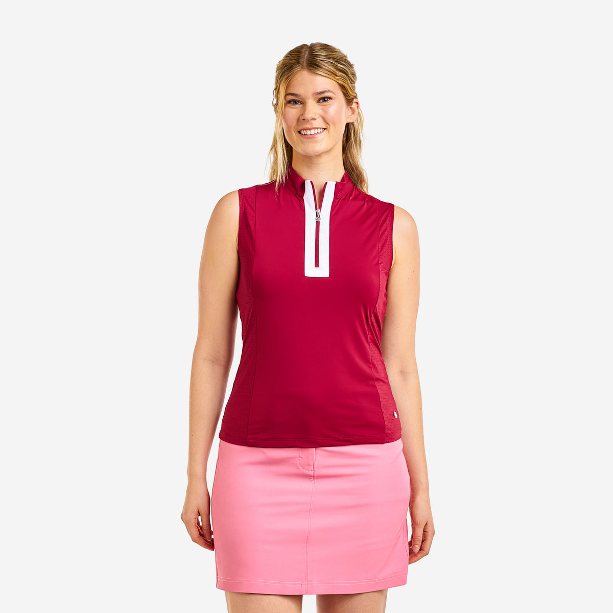 A woman modeling a sleeveless golf top in deep magenta with a white zipper placket, paired with a light pink skirt, against a solid light gray background.